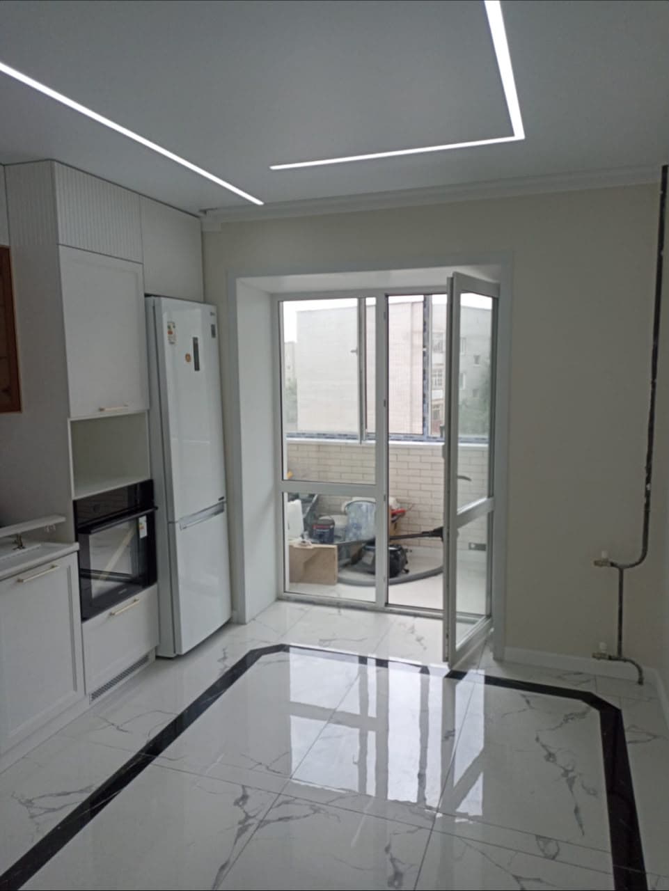 Modern white kitchen with marble floors, linear ceiling lights, and a glass balcony door.