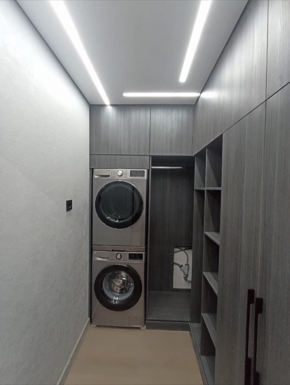 Stacked silver washer and dryer built into dark grey cabinetry with modern linear ceiling lights.