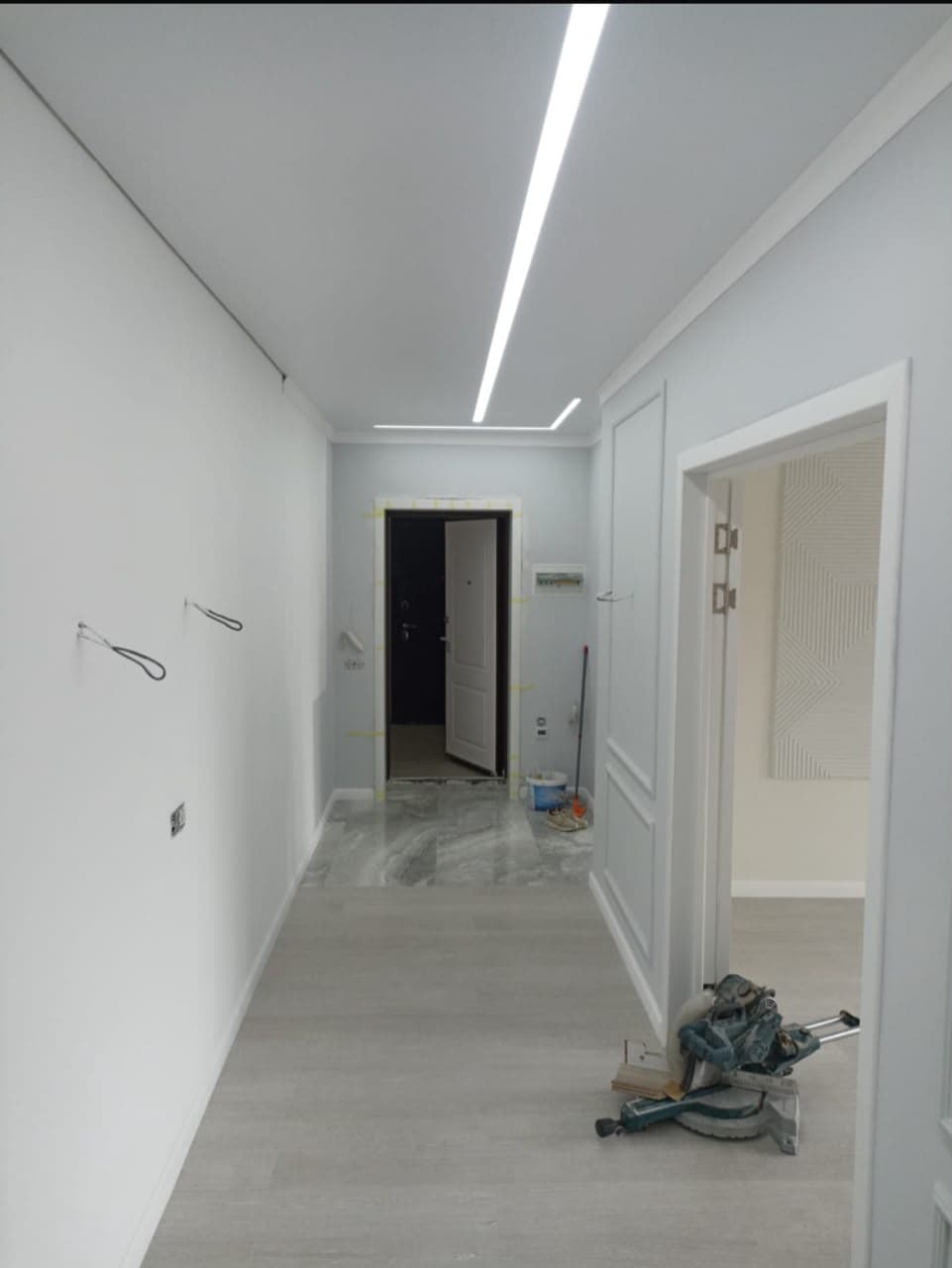 Modern hallway renovation with linear LED ceiling lighting, light grey walls, and a miter saw.
